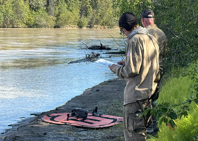 Two firefighters stand on a river bank looking at a drone sitting on an orange fabric platform.