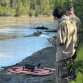 Two firefighters stand on a river bank looking at a drone sitting on an orange fabric platform.