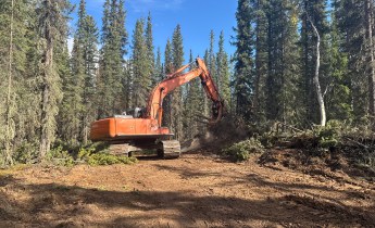 An orange tracked excavator works on a cleared fire line in a boreal forest, moving soil and woody debris during suppression-repair operations on the Mukluk Fire.