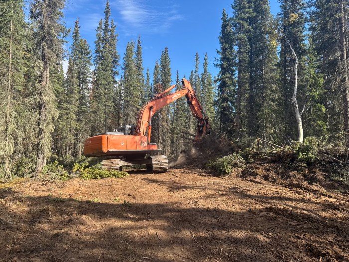 An orange tracked excavator works on a cleared fire line in a boreal forest, moving soil and woody debris during suppression-repair operations on the Mukluk Fire.