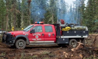 A red Willow Fire Department wildland fire engine is parked in a wooded, fire-impacted area near Tok, Alaska, with firefighters and light smoke visible in the background from pile burning.