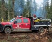 A red Willow Fire Department wildland fire engine is parked in a wooded, fire-impacted area near Tok, Alaska, with firefighters and light smoke visible in the background from pile burning.