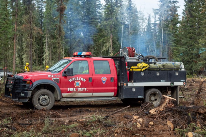 A red Willow Fire Department wildland fire engine is parked in a wooded, fire-impacted area near Tok, Alaska, with firefighters and light smoke visible in the background from pile burning.