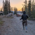 A camp crew member in a black jacket, camouflage pants, and rubber boots walks backward along a gravel road, stretching out a length of white fire hose during an active hose-rolling operation. Hose piles, rolling equipment, canopy tents, and support vehicles are visible in a forested camp at sunset, with mountains in the distance.