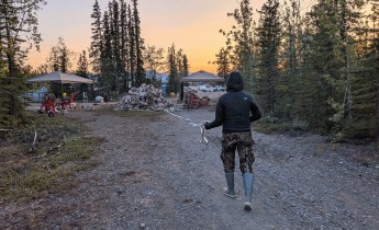A camp crew member in a black jacket, camouflage pants, and rubber boots walks backward along a gravel road, stretching out a length of white fire hose during an active hose-rolling operation. Hose piles, rolling equipment, canopy tents, and support vehicles are visible in a forested camp at sunset, with mountains in the distance.