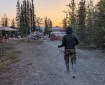 A camp crew member in a black jacket, camouflage pants, and rubber boots walks backward along a gravel road, stretching out a length of white fire hose during an active hose-rolling operation. Hose piles, rolling equipment, canopy tents, and support vehicles are visible in a forested camp at sunset, with mountains in the distance.