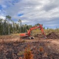 An orange excavator works beside a mound of soil and vegetation on a dozer line within the Mukluk Fire area, with charred ground and boreal forest under a partly cloudy sky.