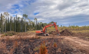An orange excavator works beside a mound of soil and vegetation on a dozer line within the Mukluk Fire area, with charred ground and boreal forest under a partly cloudy sky.