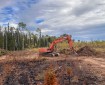 An orange excavator works beside a mound of soil and vegetation on a dozer line within the Mukluk Fire area, with charred ground and boreal forest under a partly cloudy sky.