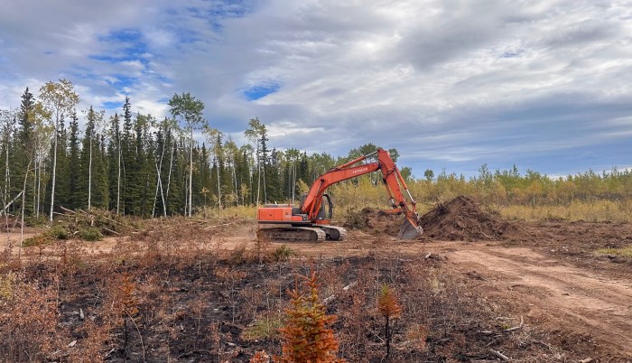 An orange excavator works beside a mound of soil and vegetation on a dozer line within the Mukluk Fire area, with charred ground and boreal forest under a partly cloudy sky.
