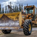 Yellow Champion road grader with a front-mounted snowplow-style blade parked on a gravel road, with a white water tender behind it and spruce trees under a cloudy sky.