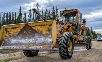 Yellow Champion road grader with a front-mounted snowplow-style blade parked on a gravel road, with a white water tender behind it and spruce trees under a cloudy sky.