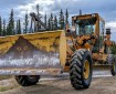 Yellow Champion road grader with a front-mounted snowplow-style blade parked on a gravel road, with a white water tender behind it and spruce trees under a cloudy sky.