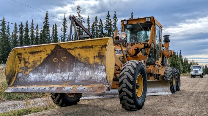 Yellow Champion road grader with a front-mounted snowplow-style blade parked on a gravel road, with a white water tender behind it and spruce trees under a cloudy sky.