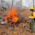 A firefighter dressed in protective gear and holding a tool watches a fire burn a debris pile.