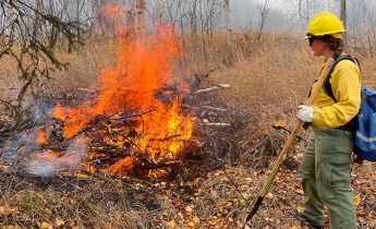 A firefighter dressed in protective gear and holding a tool watches a fire burn a debris pile.